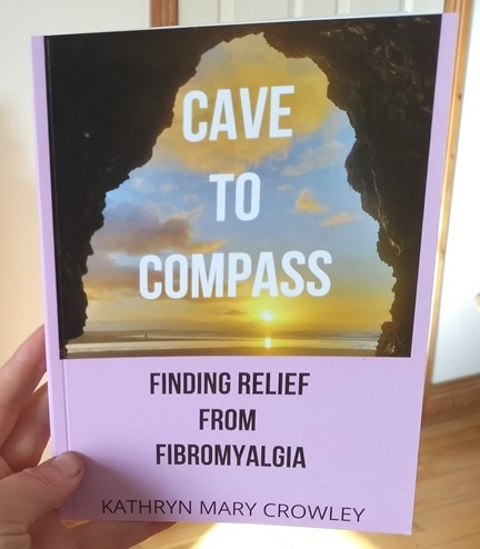 I am holding 'Cave to Compass.' My book cover shows a view of the sea from within a cave. The sun glows on the water. Half of the front cover is a lilac cover.